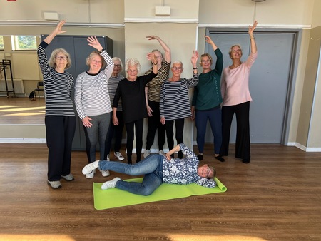 En grupp äldre kvinnor poserar glatt i ett ljust rum, en kvinna ligger på en grön yogamatta på golvet.