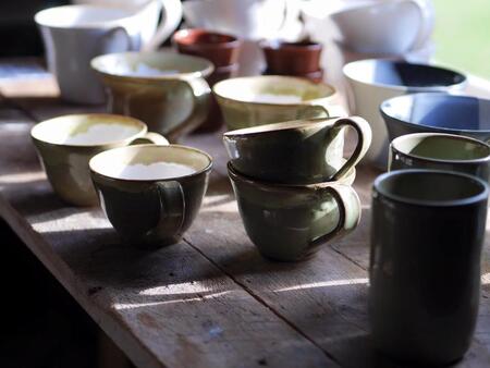 Flera keramikmuggar och koppar står på ett träbord i solljus.