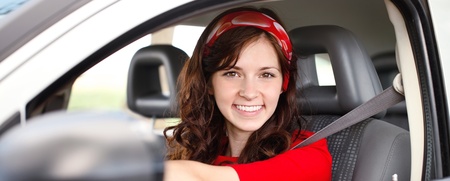 Ung kvinna med långt brunt hår och röd tröja sitter leende bakom ratten i en bil.