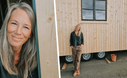 Person med långt grått hår står framför en träbyggnad med hjul och en kruka på marken.