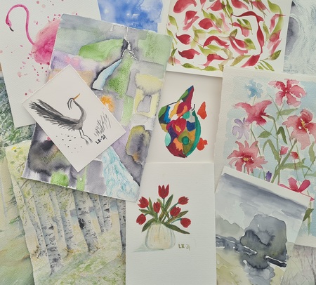 Flera akvarellmålningar med blommor, fåglar och naturmotiv ligger utspridda på ett bord.