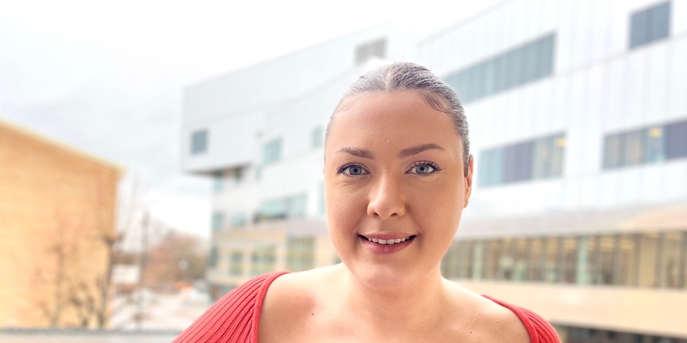 Kvinna i röd tröja står på en balkong med moderna byggnader i bakgrunden.