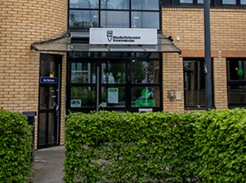 Entré till en modern byggnad med ljusgul tegelfasad, stora fönster och glasparti i mitten.