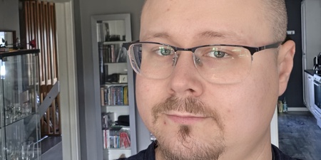 Man med glasögon och kort skägg tar en selfie i ett modernt vardagsrum med grå väggar.