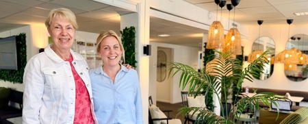 Två personer står tillsammans i ett modernt, ljust kontorslandskap med gröna växter och soffor.
