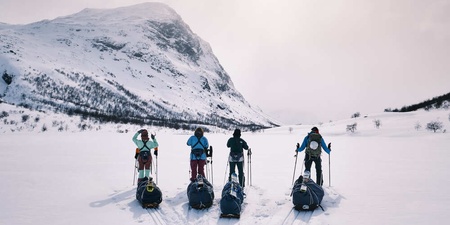 Fem personer med ryggsäckar och pulkor står på skidor i ett snötäckt fjällandskap med berg i bakgrunden.