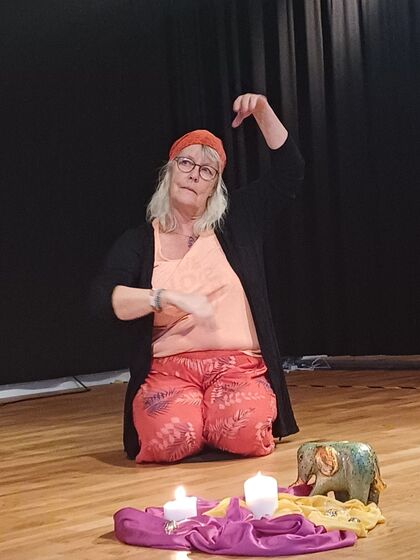 Kvinna med glasögon och röd sjal på huvudet sitter på knä och gestikulerar på en scen med tända ljus och tyg framför sig.