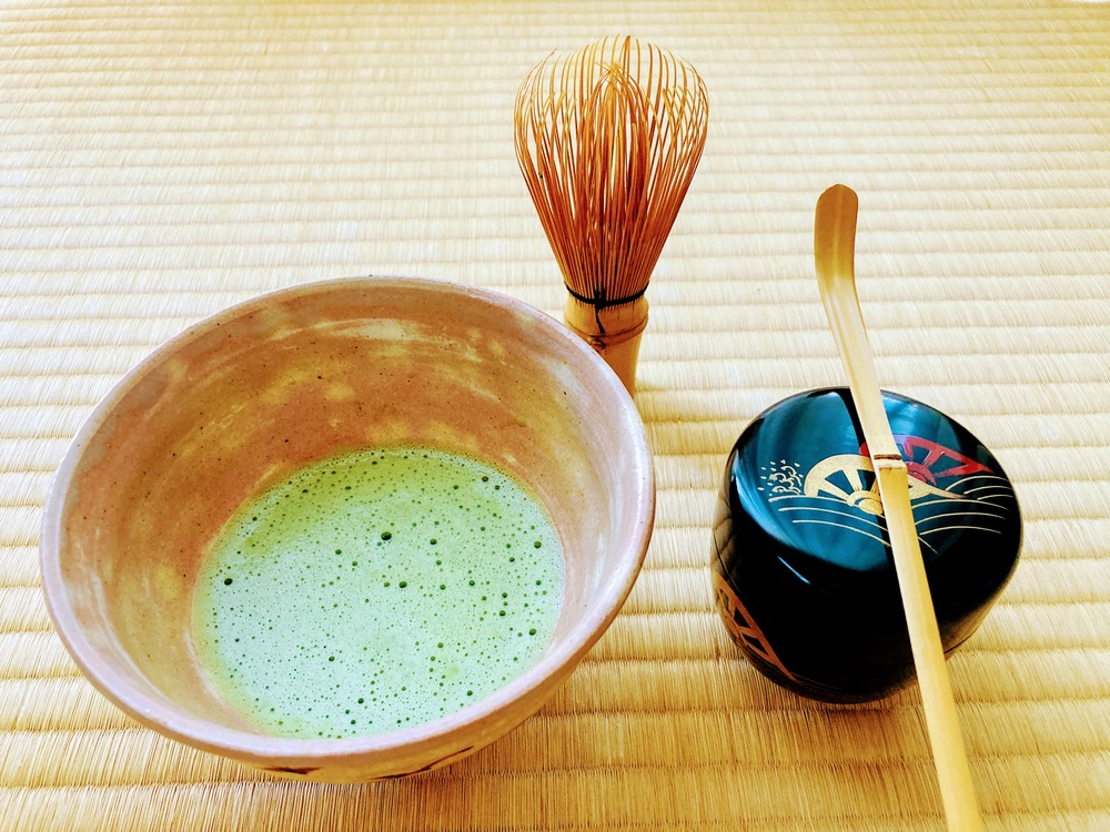 En skål med matcha-te, en bambuvisp, en lackad burk och en bambusked på tatamimatta.