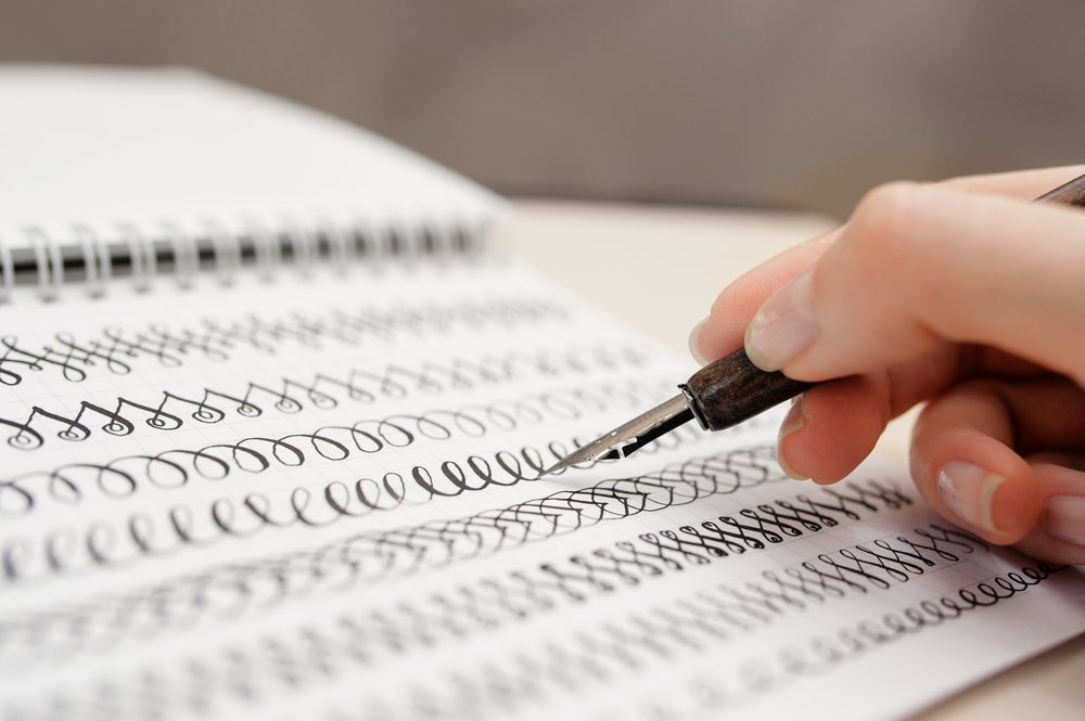 En hand övar kalligrafi med en bläckpenna i en anteckningsbok fylld med övningsslingor.