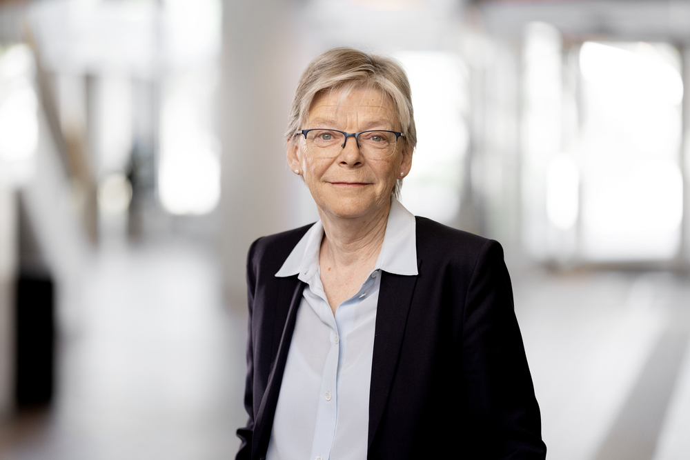 Äldre kvinna med glasögon och kort hår i kostym står inomhus i ljus, suddig miljö.
