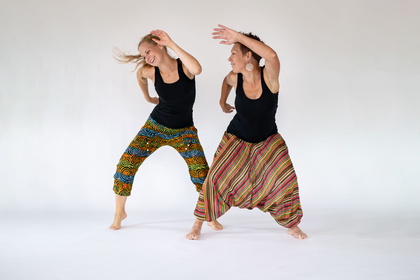 Två kvinnor dansar glatt i färgglada byxor mot en vit bakgrund.