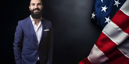 Man i blå kostym står bredvid en amerikansk flagga mot mörk bakgrund.