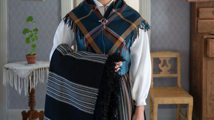 Person i traditionell folkdräkt håller en svart filt, inomhus med gammaldags inredning.