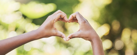 Två händer formar ett hjärta mot en suddig, grön bakgrund utomhus.