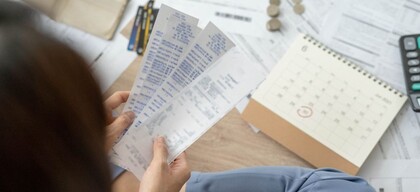 En person håller flera kvitton framför sig, omgiven av papper, mynt, en kalender och en miniräknare.