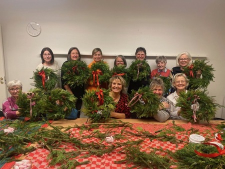 En grupp glada personer visar upp egentillverkade julkransar vid ett bord täckt med granris och dekorationer.