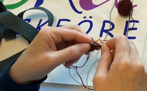 Två händer stickar med garn och stickor ovanpå ett papper med färgglad text.