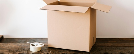 En öppen kartong står på ett träbord bredvid en sax och en rulle tejp.