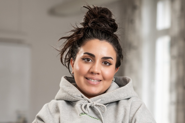 Kvinna med mörkt hår i knut och grå hoodie står med armarna i kors i ett klassrum.