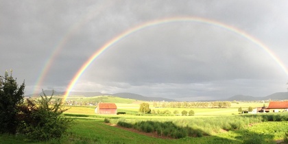 Landskap med gröna fält, bondgårdar och en tydlig dubbel regnbåge mot en molnig himmel.