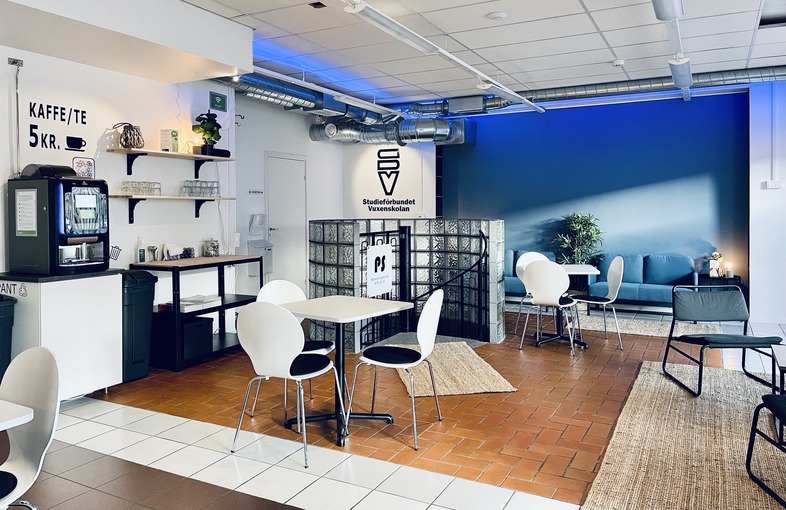 Ett modernt café med vita stolar, små bord, kaffemaskin och blå vägg. Inga människor syns i lokalen.