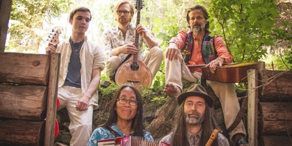 Fem personer med musikinstrument sitter utomhus i en grönskande skogsmiljö.