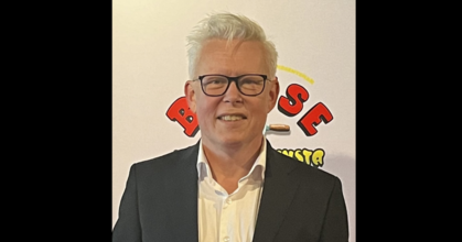 Man med vitt hår och glasögon i kostym står framför en ljus bakgrund med färgglad text.