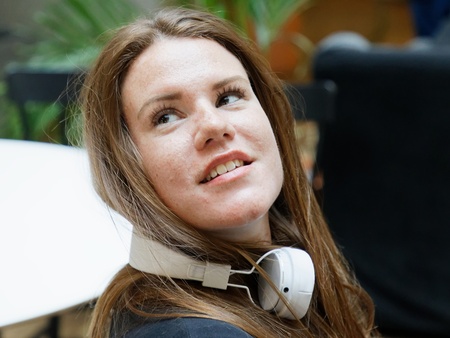 Kvinna med långt brunt hår och hörlurar runt halsen sitter inomhus och tittar uppåt.