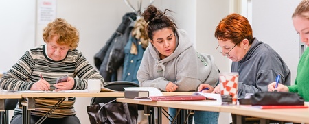Fyra personer sitter vid ett bord och arbetar tillsammans med papper och pennor i ett klassrum.