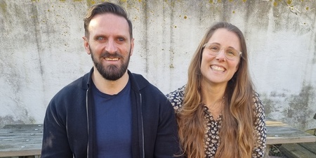 Två personer sitter på en bänk utomhus med en svart ledarhund mellan sig.