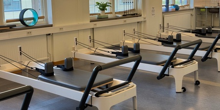 Ett ljust pilatesrum med flera reformermaskiner, stora fönster och träningsredskap på fönsterbrädorna.