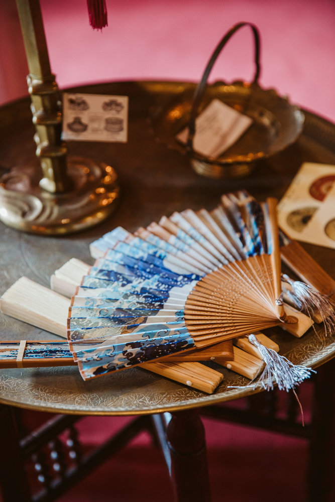 En dekorativ solfjäder med blått mönster ligger på ett runt bord av metall tillsammans med andra små föremål.