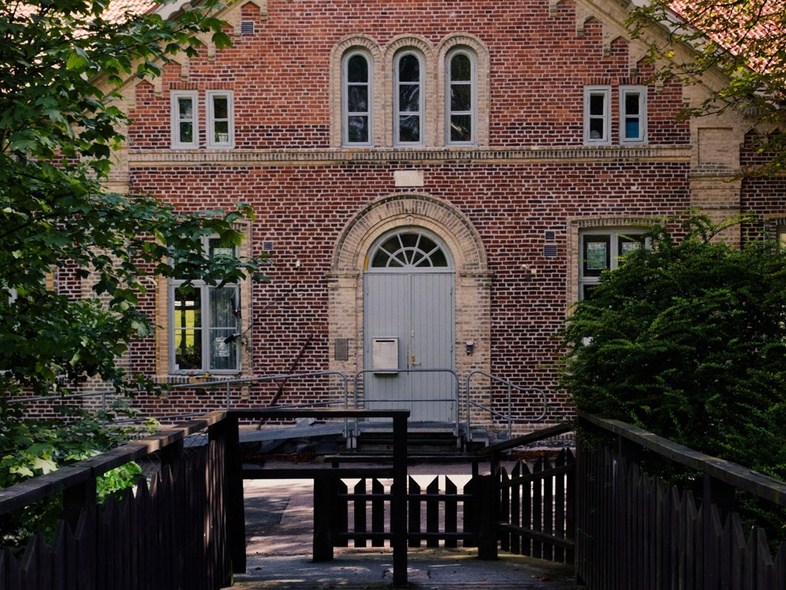 En gammal tegelbyggnad med spetsigt tak, sedd från en träbro omgiven av grönska.