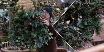 Person håller en stor grön krans av granris och blad i en rustik miljö med naturmaterial.