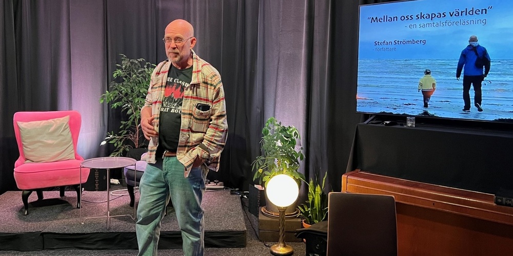 Man står på en scen med svart bakgrund, bredvid en TV-skärm med text och en bild på två personer vid havet.