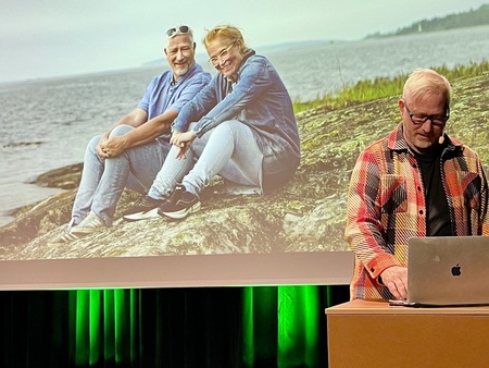 Person i rutig skjorta står vid ett podium med en dator som visar en stor bild på projektorduk i bakgrunden. På den bilden är det två personer som sitter på en sten vid vatten.