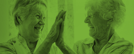 Två äldre kvinnor skrattar och ger varandra en high five i ett ljust rum.