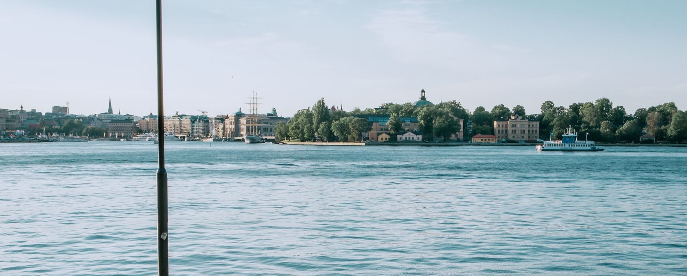 Två personer sitter vid vattnet på en kaj med utsikt över en stad och båtar i bakgrunden.