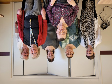 Sex kvinnor, varav en sitter i rullstol, poserar tillsammans inomhus framför en vit vägg.