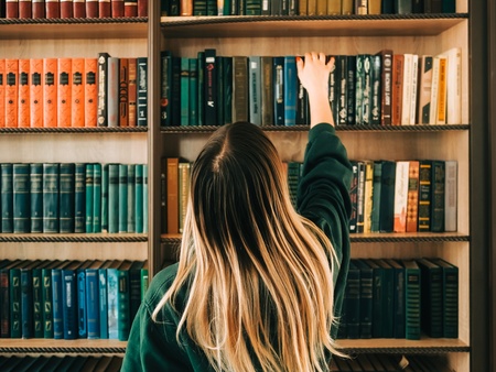 En person med långt blont hår sträcker sig efter en bok på en bokhylla fylld med böcker.