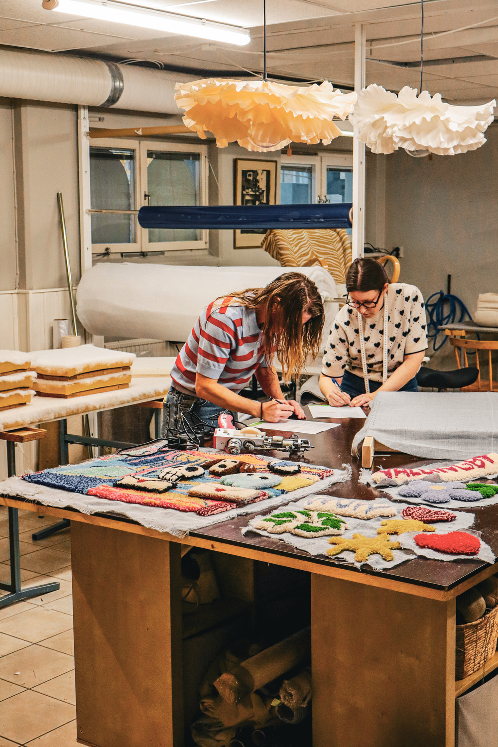Två personer arbetar med textilkonst i en färgglad ateljé med tygprover och lampor i taket.