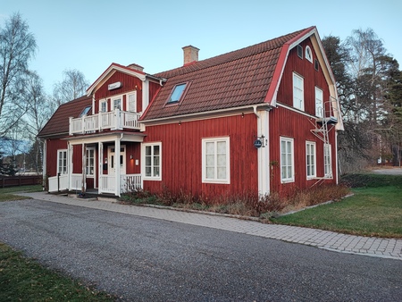 Rött trähus med vita knutar och tegeltak, omgiven av gräsmatta och träd, en lugn landsbygdsmiljö.
