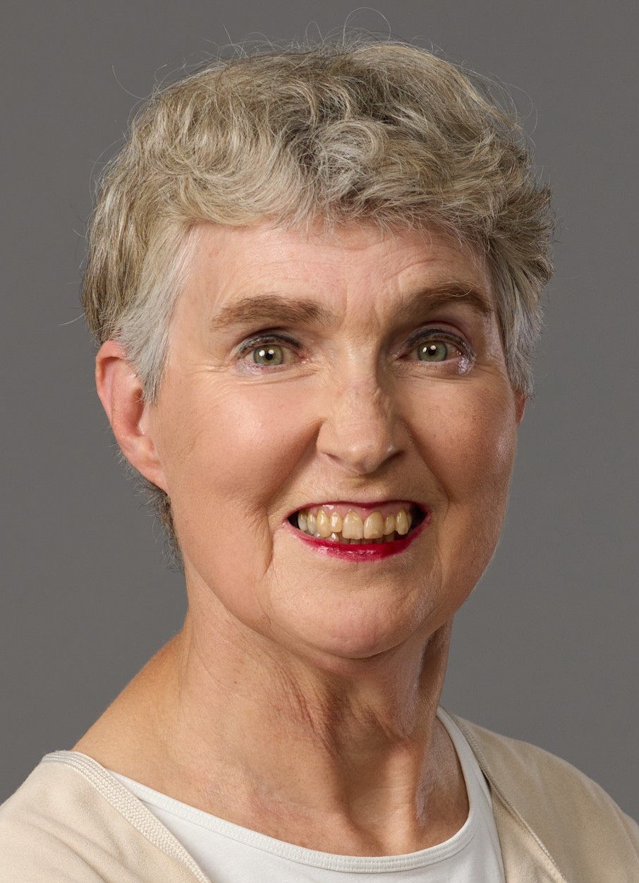 Äldre person med grått, lockigt hår och ljus tröja mot grå bakgrund.