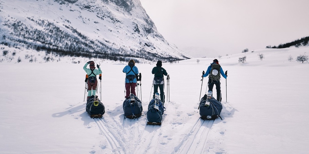 Fyra personer med pulkor står på skidor i ett snötäckt fjällandskap med berg i bakgrunden.