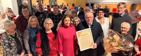 En grupp människor står samlade inomhus, en person håller ett diplom och en annan en blomsterbukett.