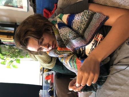 Person med glasögon sitter i soffa och stickar, insvept i en färgglad stickad halsduk.