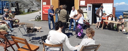 En grupp människor samlade utomhus vid en röd byggnad, några sitter på stolar och andra står och pratar.