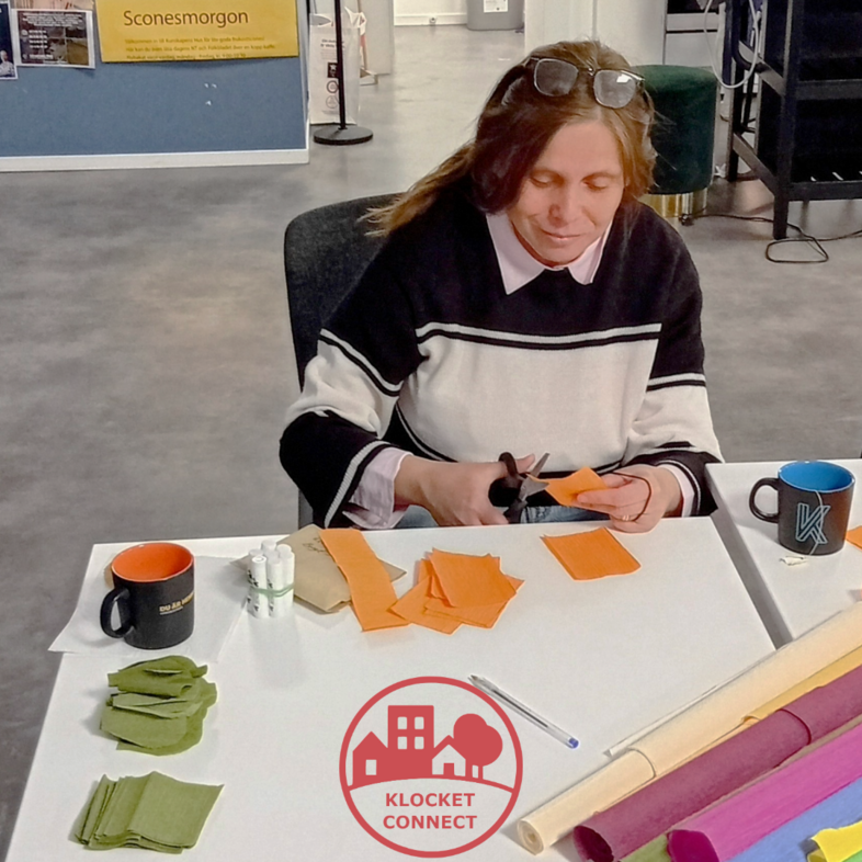 En kvinna sitter vid ett bord och klipper färgat papper, med muggar och pysselmaterial framför sig.