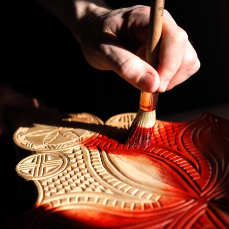 En hand målar röd färg på ett träsnideri med dekorativa mönster.