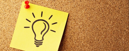 Gul post-it-lapp med en tecknad glödlampa, fäst med en röd nål på en anslagstavla av kork.